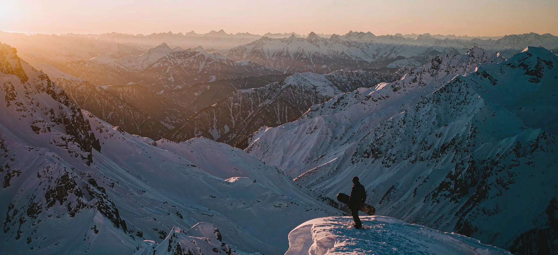 A snowboarder silhouetted on a ridge at sunrise above an alpine panorama