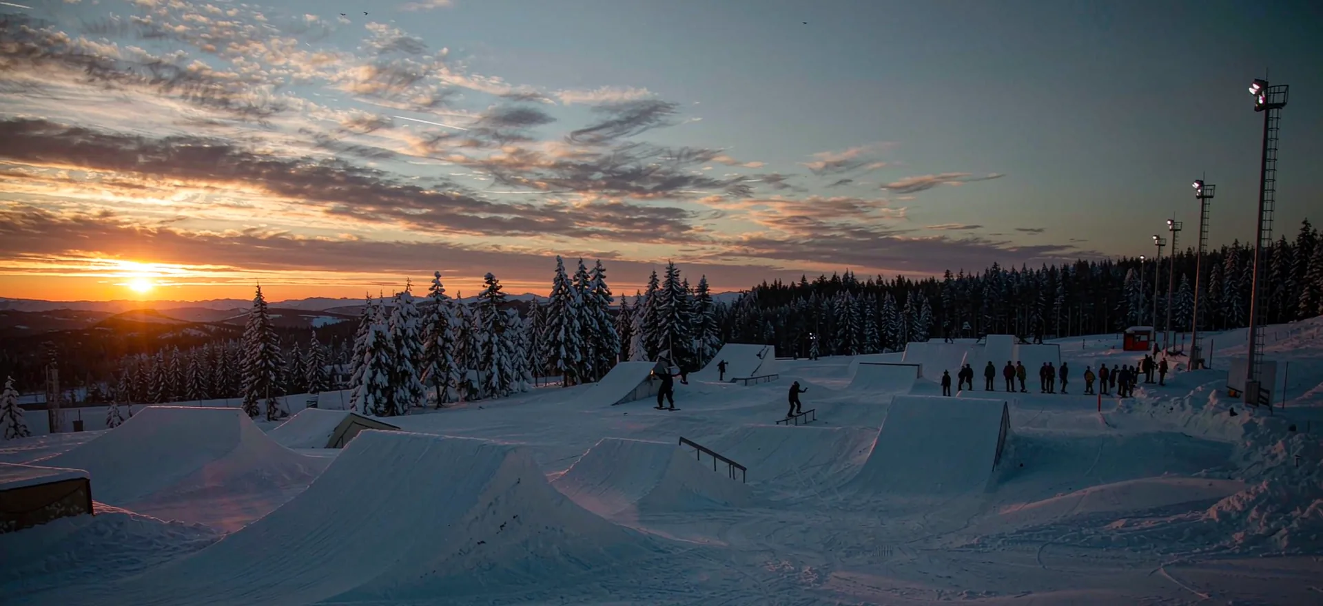 A snow park at sunset with riders queuing at the drop-in