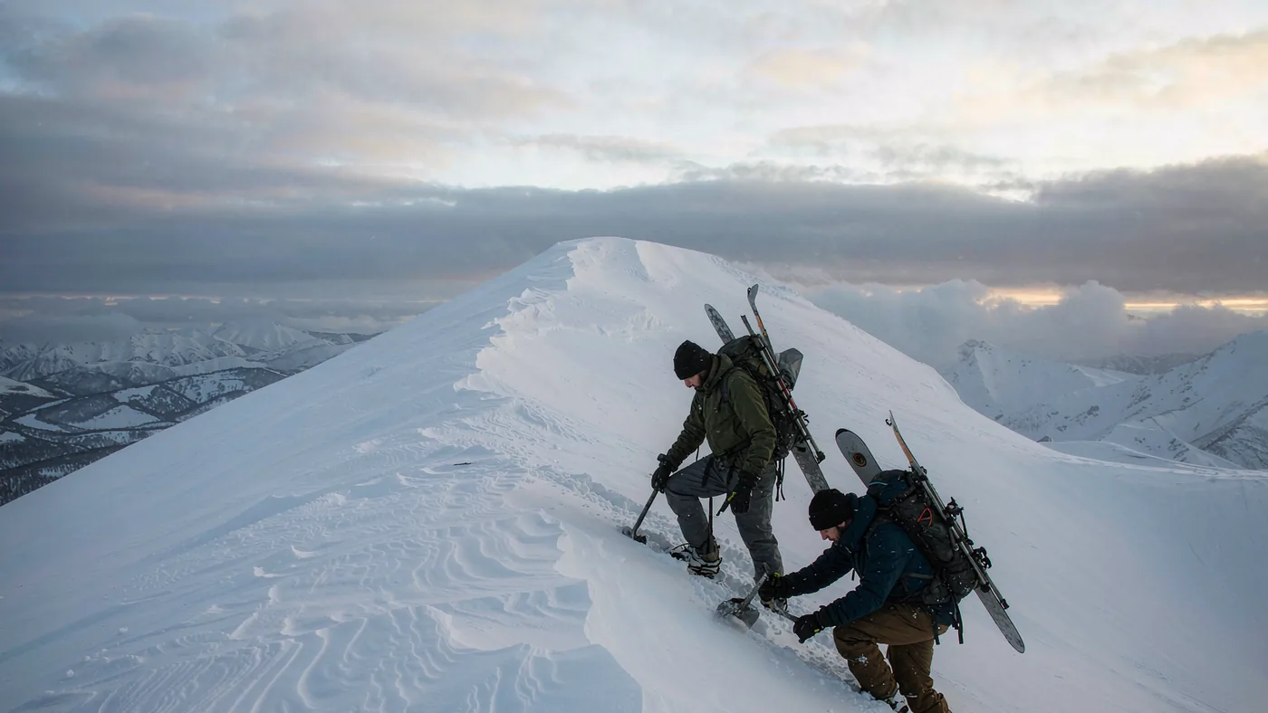 Two splitboarders with packs and shovels reading a slope on a wind-affected ridge