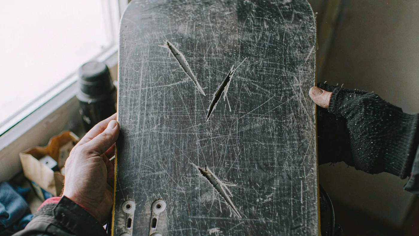 Hands running along the base of a used snowboard held up to window light to reveal scratches